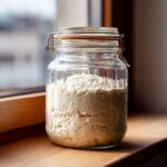 A bubbly, active sourdough starter filling a clear glass jar resting on a wooden windowsill.