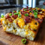 A close-up slice of baked potato casserole topped with melted cheddar cheese, crispy bacon, and green onions.
