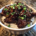 A close-up of a delicious chicken rice bowl featuring dark, glazed chicken pieces served over white rice and garnished with sliced green onions and sesame seeds.