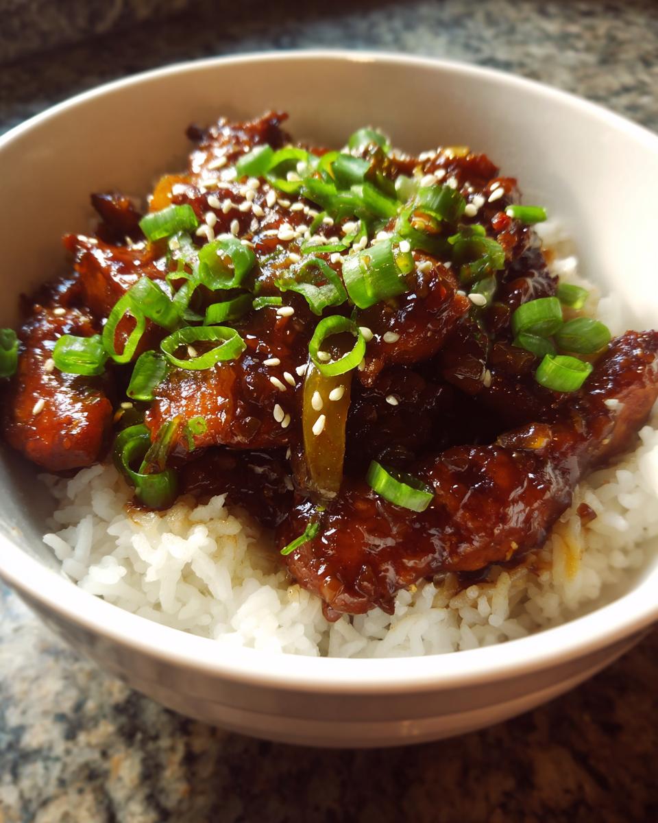 Close-up of a delicious chicken rice bowl featuring sticky, glazed chicken pieces topped with sliced green onions and sesame seeds.
