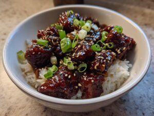 Close-up of a white bowl filled with white rice topped with glazed chicken pieces, sesame seeds, and sliced green onions for a chicken rice bowl.