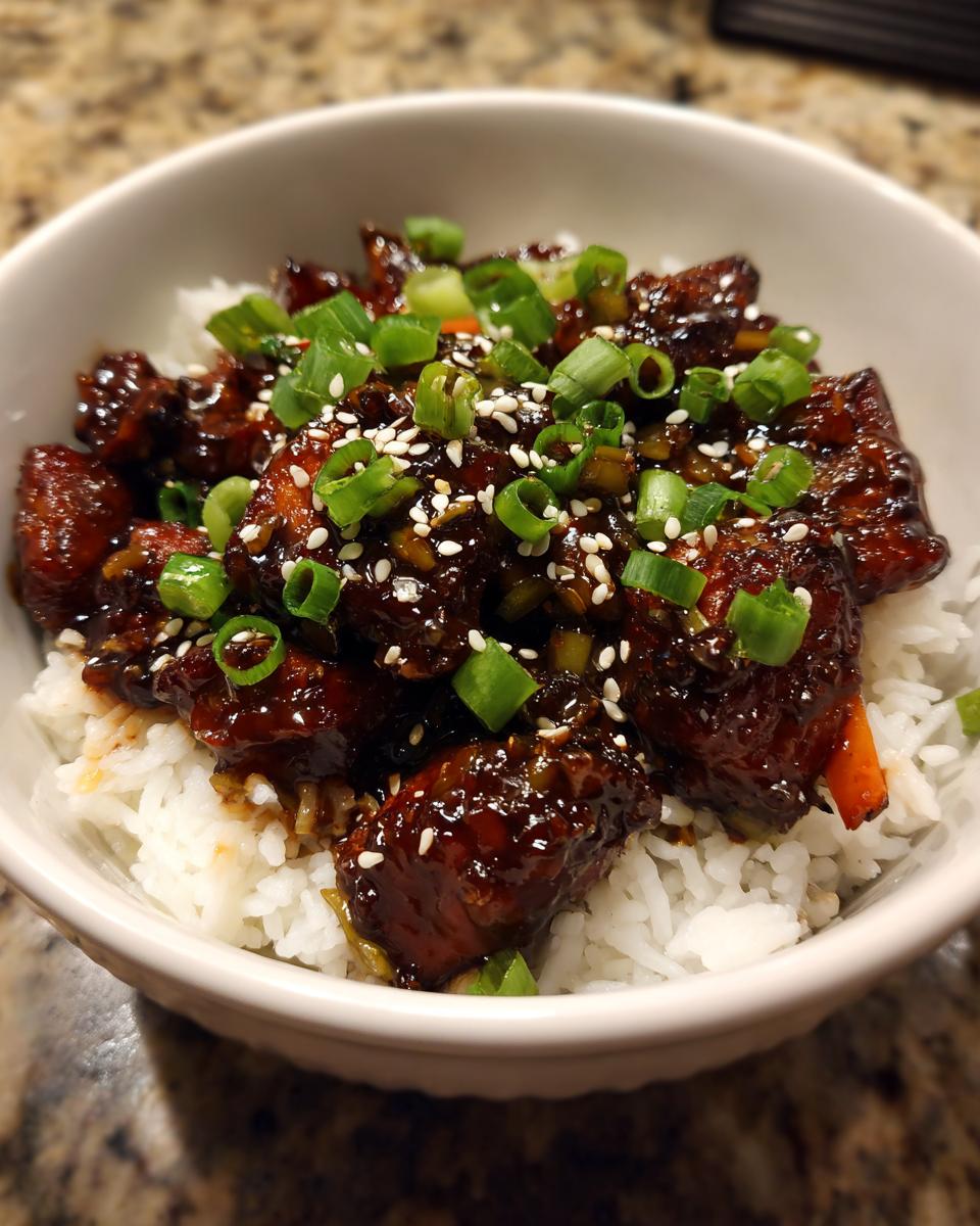 Close-up of a homemade chicken rice bowl featuring dark, glazed chicken pieces over white rice, garnished with sesame seeds and sliced green onions.