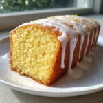 A close-up of a moist, golden slice from a lemon cake recipe loaf drizzled with white glaze and lemon zest.