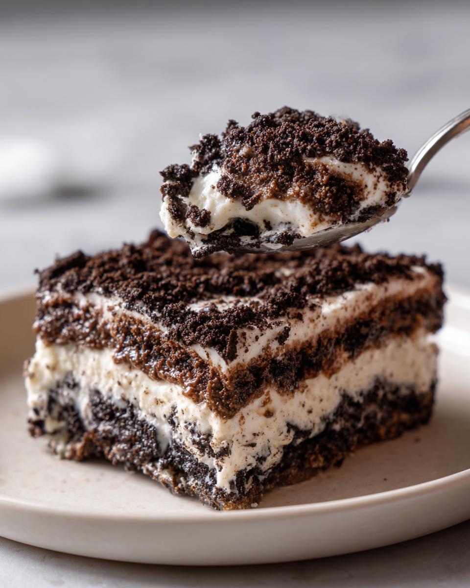 A spoonful of creamy, layered oreo dirt cake being lifted above a square serving on a plate.