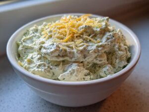 Close-up of creamy, dill-flecked pickle dip topped with shredded cheddar cheese in a white bowl.
