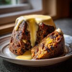 A rich, dark plum pudding, partially sliced, generously covered in flowing vanilla custard sauce.