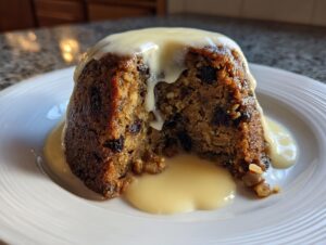 A cross-section of a rich, dark plum pudding topped with thick, flowing custard sauce on a white plate.