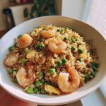 Close-up of a white bowl filled with flavorful shrimp fried rice, featuring plump shrimp and bright green peas.