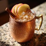 A frosty copper mug filled with an apple cider moscow mule, garnished with apple slices and a cinnamon stick.
