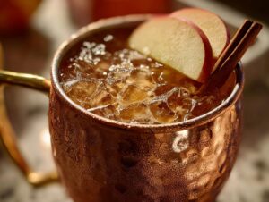 Close-up of a frosty copper mug filled with an apple cider moscow mule, garnished with apple slices and a cinnamon stick.