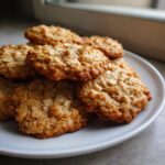A stack of freshly baked, golden brown banana oatmeal cookies resting on a white plate near a window.