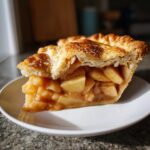 A close-up of a perfect slice of apple pie showing thick, flaky crust and generous cinnamon apple filling.