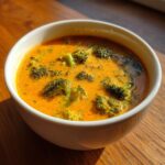 Close-up of a white bowl filled with rich, orange broccoli cheddar soup featuring visible florets of green broccoli.