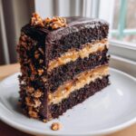 A tall slice of chocolate caramel toffee crunch cake showing three dark chocolate layers and thick caramel filling.
