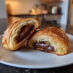 A flaky, golden-brown chocolate croissant cut in half, revealing melted chocolate filling.