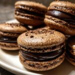 A close-up stack of decadent, dark brown chocolate macarons filled with glossy chocolate ganache.