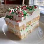 A cross-section slice of Christmas tree cheesecake featuring layers of sponge cake, creamy filling with red and green sprinkles, topped with white frosting.