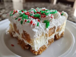 Close-up of a slice of Christmas Tree Cheesecake featuring layers of graham cracker and cream cheese filling, topped with whipped cream and red/green sprinkles.