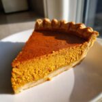 A close-up, sunlit slice of classic pumpkin pie recipe showing the rich orange filling and crimped crust edge on a white plate.
