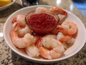 A white bowl filled with peeled, cooked shrimp surrounding a small glass ramekin of red shrimp cocktail sauce.