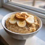 A white bowl filled with creamy banana oatmeal, topped with fresh banana slices and a dollop of peanut butter.