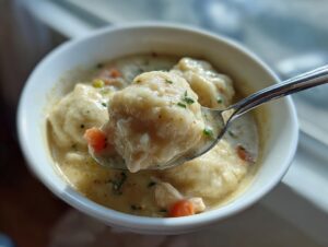 A spoonful of creamy chicken and dumpling, showing a large, soft dumpling and pieces of carrot in a white bowl.