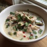 Close-up of a white bowl filled with creamy chicken coconut soup, garnished with fresh cilantro and red chili flakes.