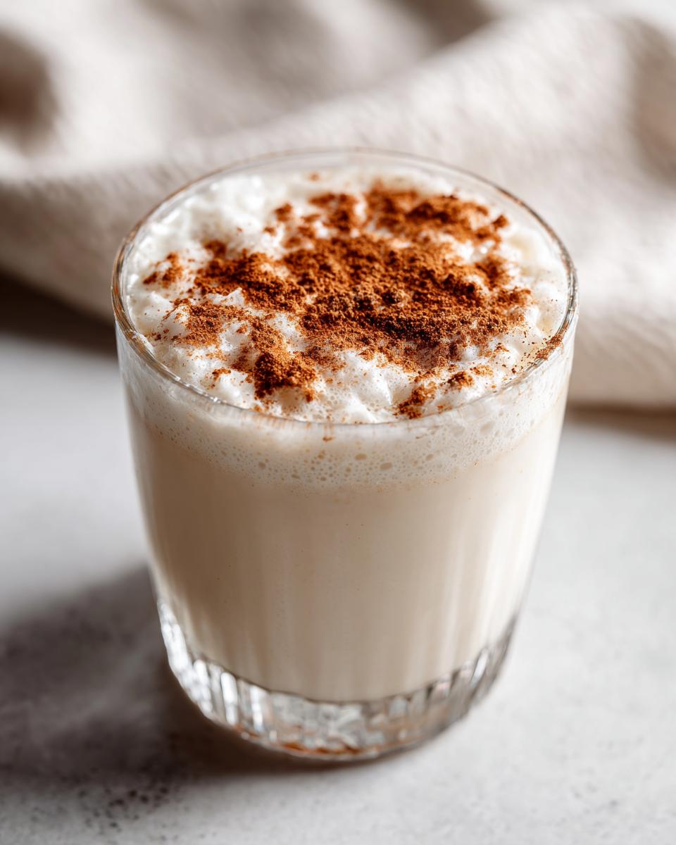 Close-up of a glass filled with creamy coquito recipe, topped with a generous dusting of ground cinnamon.