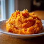 A close-up of a generous serving of bright orange, creamy mashed sweet potatoes mounded on a white plate.