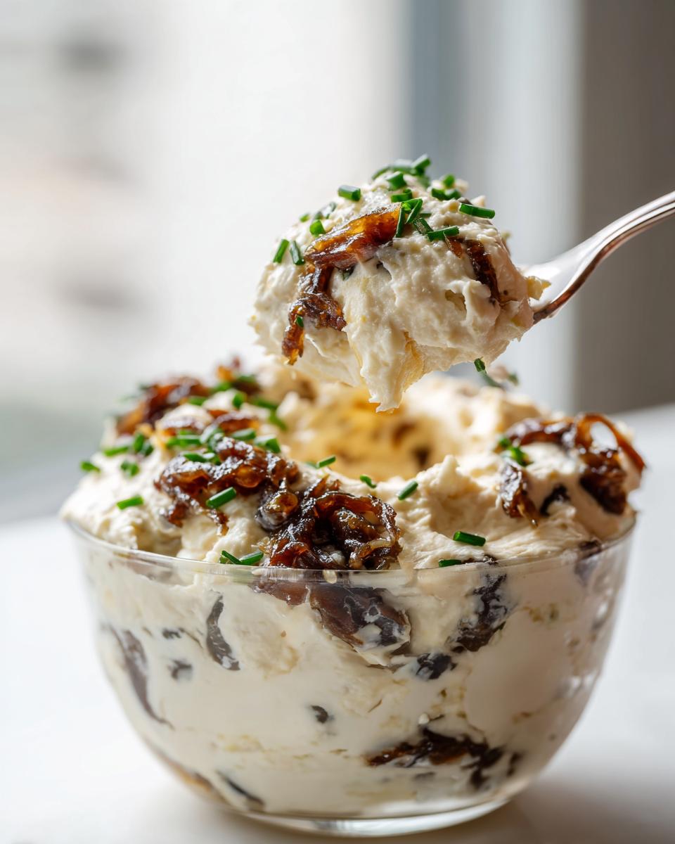 A spoonful of rich onion dip topped with caramelized onions and chives being lifted from a clear bowl.