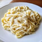 Close-up of fettuccine pasta coated heavily in rich, creamy olive garden alfredo sauce on a white plate.