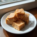 A stack of creamy, light brown penuche fudge squares piled on a small white plate near a window.
