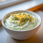 A white bowl filled with creamy green pickle dip, topped with shredded cheddar cheese and fresh dill.