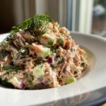 A mound of creamy salmon salad mixed with chopped celery and red onion, topped with fresh dill.