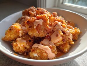 Close-up of crispy, golden brown Bang Bang Chicken pieces drizzled generously with a creamy, orange-hued sauce in a white bowl.