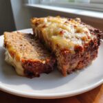 Two thick slices of moist crockpot French onion meatloaf topped with bubbly, melted cheese on a white plate.