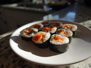 A white plate holds eight slices of homemade tuna kimbap, featuring rice, seaweed, and a bright orange tuna filling.