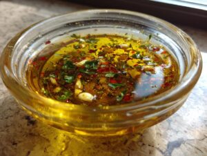 Close-up of a clear glass bowl filled with olive oil bread dip, featuring garlic, herbs, and red pepper flakes.