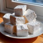 A stack of thick, square homemade marshmallows generously dusted with white powdered sugar on a plate.