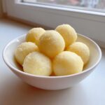 A white bowl filled with several fluffy, pale yellow potato dumplings lightly dusted with fine crumbs.