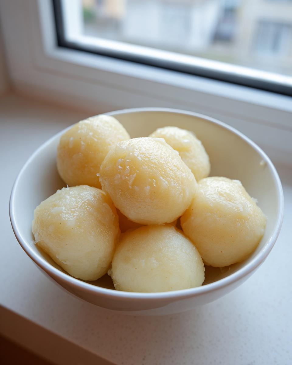 A white bowl filled with several freshly cooked, glistening potato dumplings sitting on a windowsill.