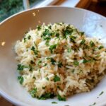 Close-up of fluffy rice pilaf served in a white bowl and garnished generously with chopped fresh parsley.