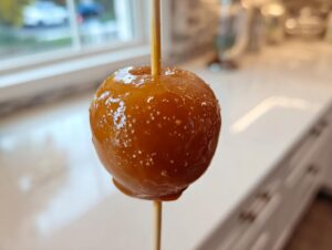 A close-up, featured image of a single, glossy caramel apple on a wooden stick, ready to eat.