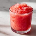 Close-up of a frosty, bright red strawberry daiquiri served in a clear glass on a white marble surface.
