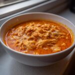 Close-up of a white bowl filled with creamy, orange-red buffalo chicken chili featuring shredded chicken and white beans.