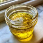 Close-up of golden homemade salad dressing with herbs and spices settling in a small glass jar.