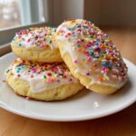 Three soft Italian ricotta cookies topped with white glaze and colorful sprinkles stacked on a white plate.