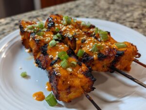 Close-up of three grilled bang bang chicken skewers drizzled with orange sauce, sesame seeds, and green onions.