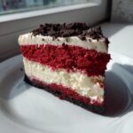 A tall, layered slice of red velvet cheesecake featuring alternating red cake and white cream cheese layers, topped with dark crumbs.