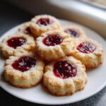 A close-up of several light-colored thumbprint cookies topped with bright red jam and sprinkled with sugar.